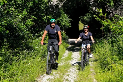 un uomo ed una donna in mountain bike in una stradina nel verde in uscita da un sottopassaggio
